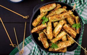 A bowl of golden-brown, crispy zucchini fries garnished with parsley, surrounded by bamboo skewers and a checkered cloth.