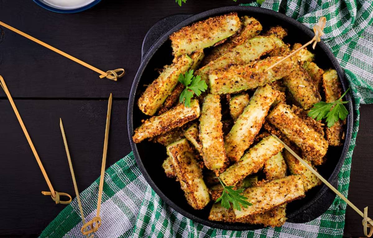 A bowl of golden-brown, crispy zucchini fries garnished with parsley, surrounded by bamboo skewers and a checkered cloth.