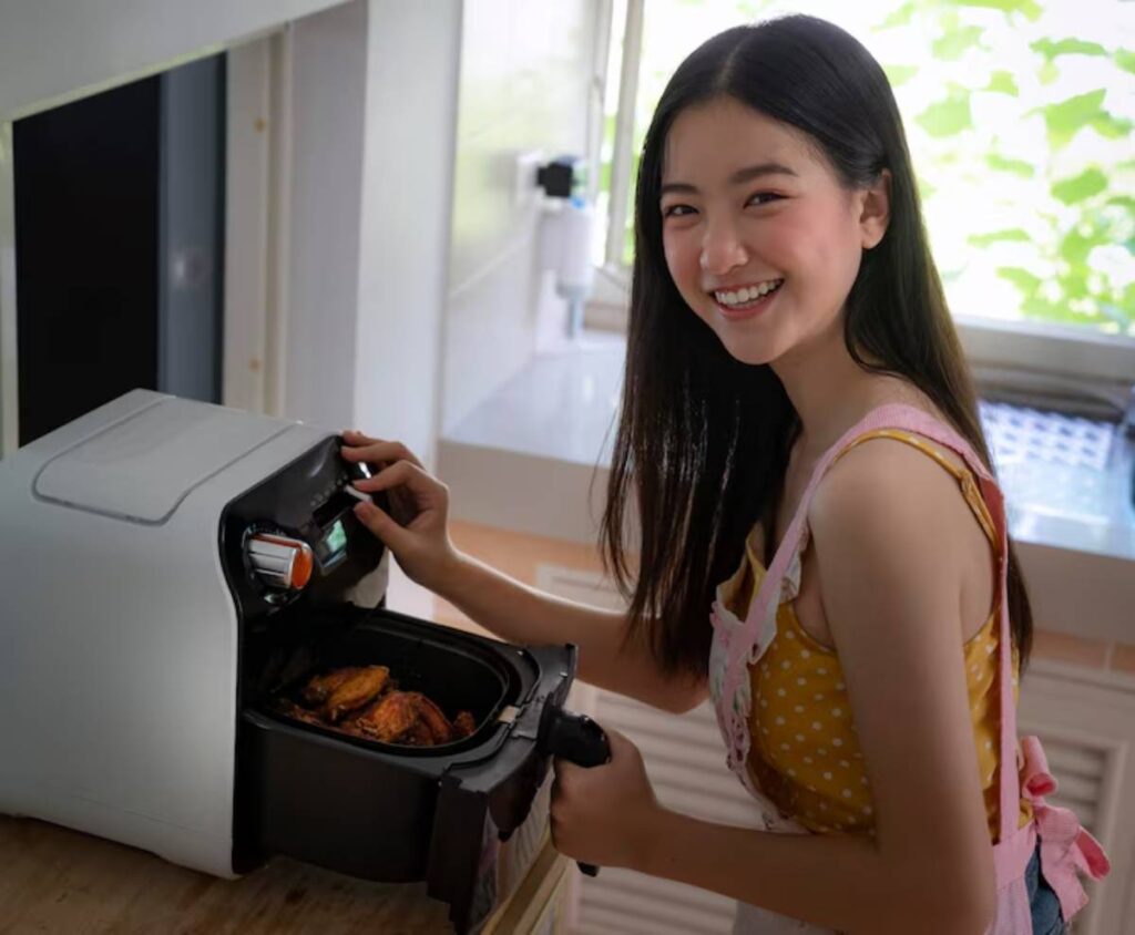 A person wearing a yellow polka-dot top and pink apron reaches into an air fryer filled with golden-brown cooked food in a bright kitchen.