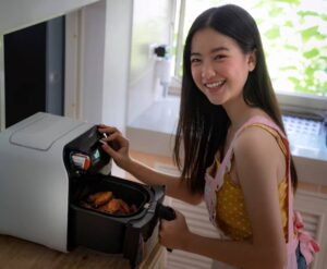 A person wearing a yellow polka-dot top and pink apron reaches into an air fryer filled with golden-brown cooked food in a bright kitchen.