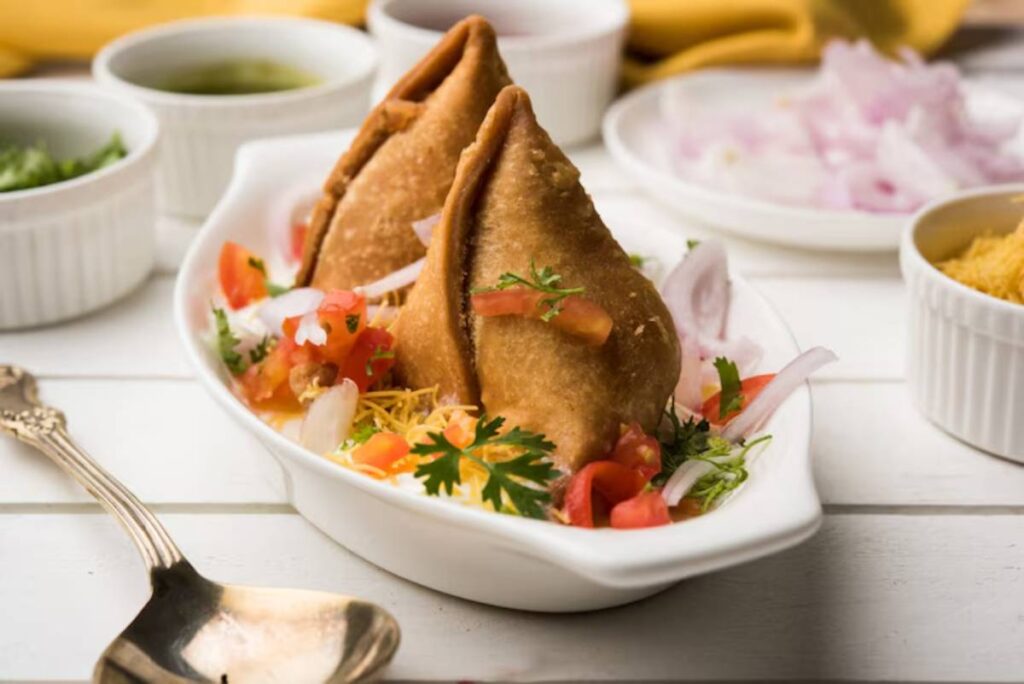 Two crispy samosas topped with diced tomatoes, onions, and cilantro, served with sauces in bowls on a rustic wooden table.
