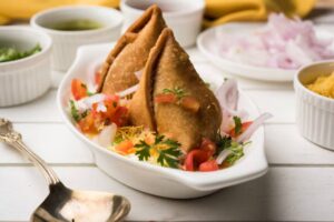 Two crispy samosas topped with diced tomatoes, onions, and cilantro, served with sauces in bowls on a rustic wooden table.