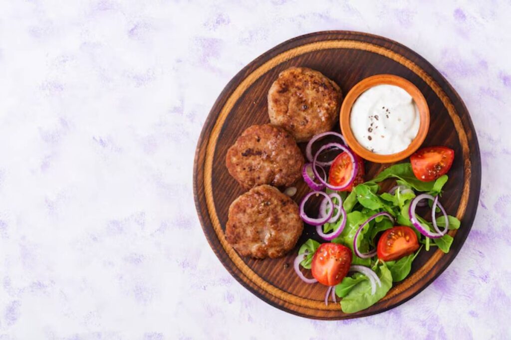 A wooden plate holds three meat patties, fresh salad with cherry tomatoes, and a bowl of creamy sauce, all against a light background.