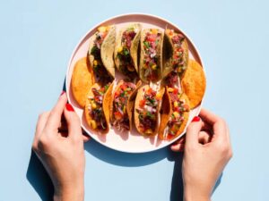 A plate of colorful tacos filled with meat, vegetables, and salsa, held by hands with red nail polish, against a light blue background.