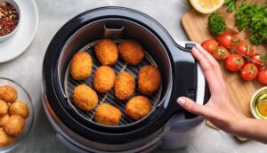 A hand reaches for golden-brown croquettes inside an air fryer, surrounded by fresh tomatoes, herbs, and spices on a kitchen counter.